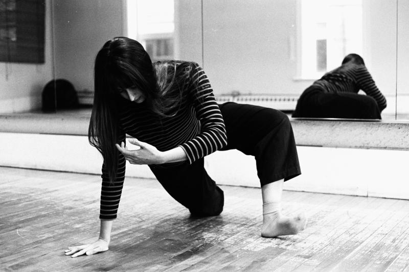A dancer in black pants and a striped shirt step plants one foot and one hand from a kneeling position