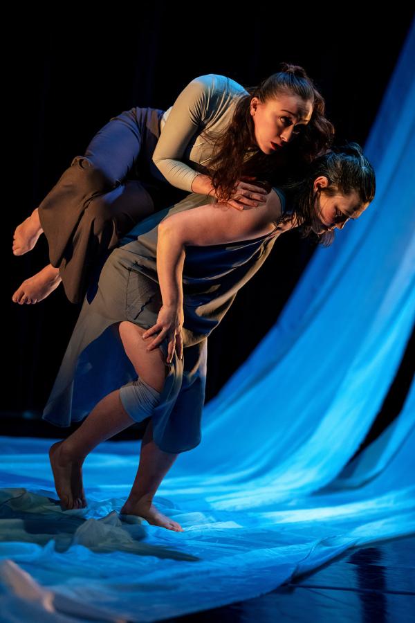 A settler white woman carries an Indigenous woman on her back across white fabric in a blue light. 