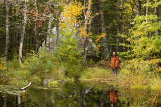 A woman stands in a forest next to a pond.