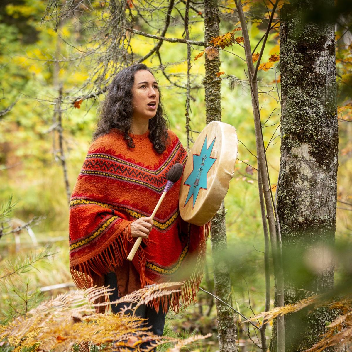 A woman stands in a forest holding a drum