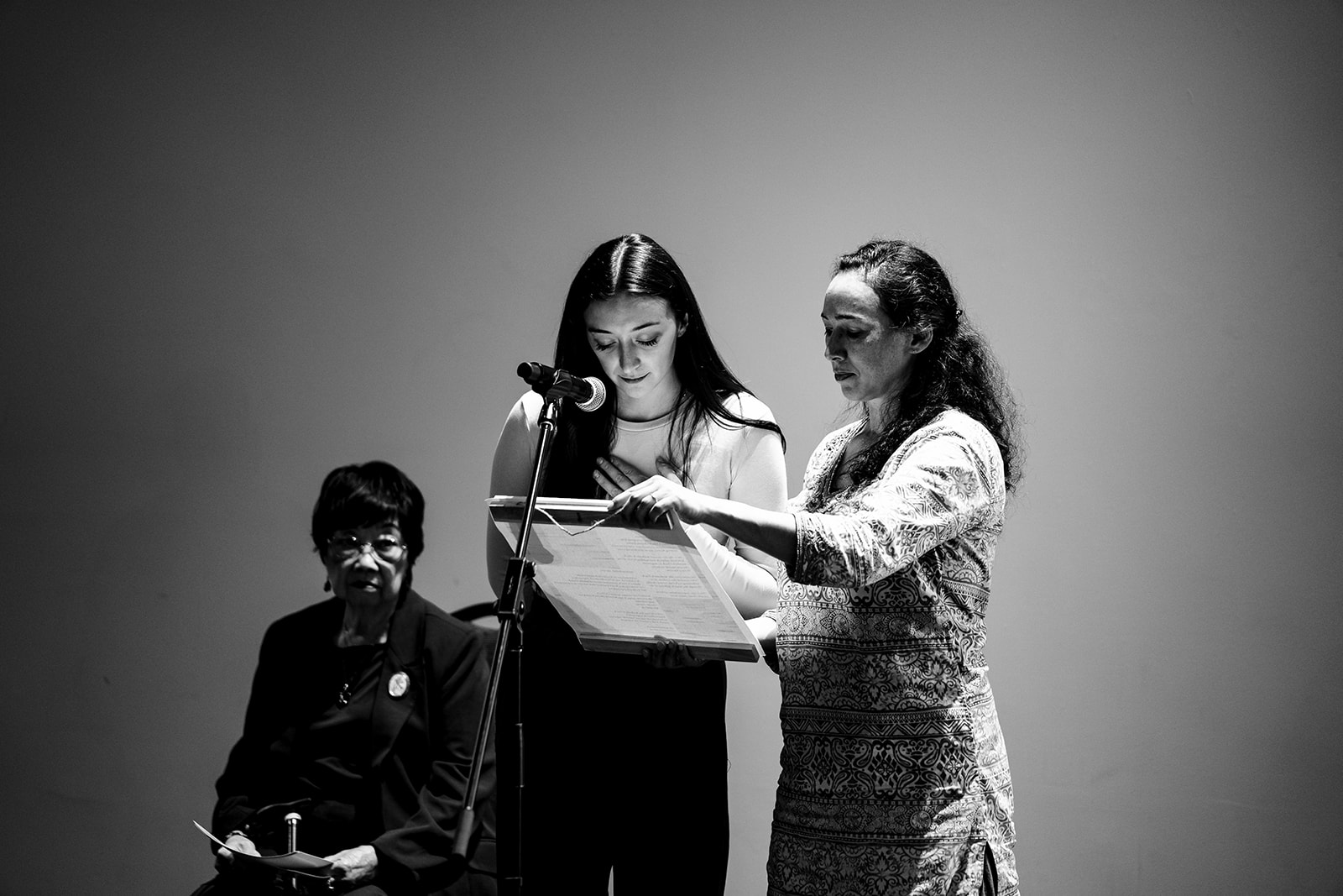 Two people stand in front of a mic and read from a scroll of paper