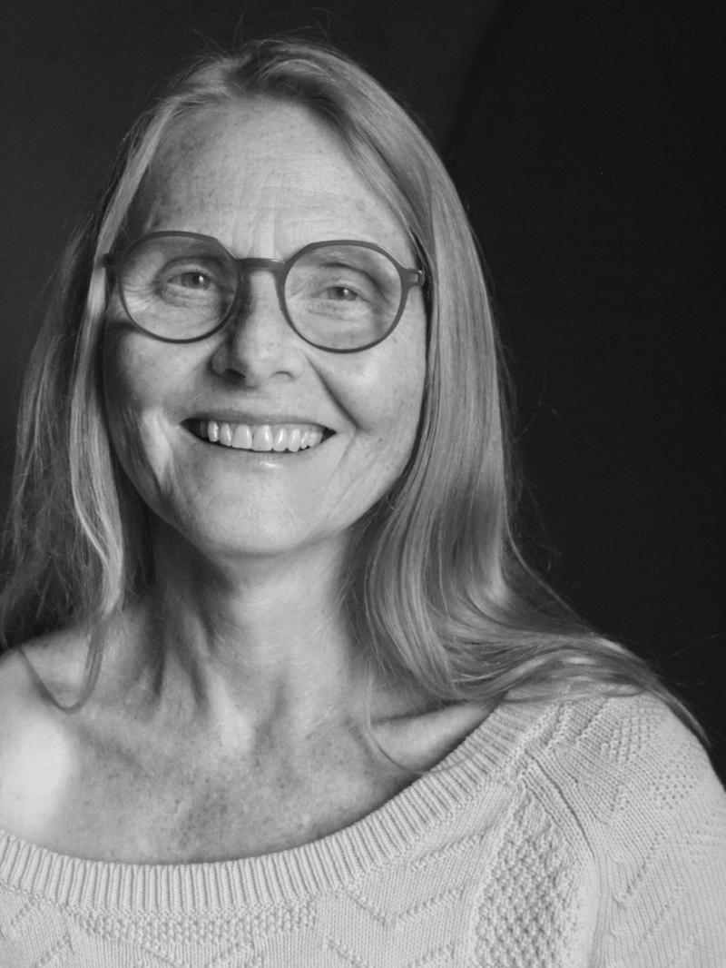 Black and white photo of a smiling woman with long light hair and glasses, wearing a light knitted shirt off the right shoulder