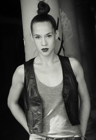 Black and white image of a light-skinned woman with dark red lipstick, and hair in a high bun. She is wearing a light tank top and dark vest.