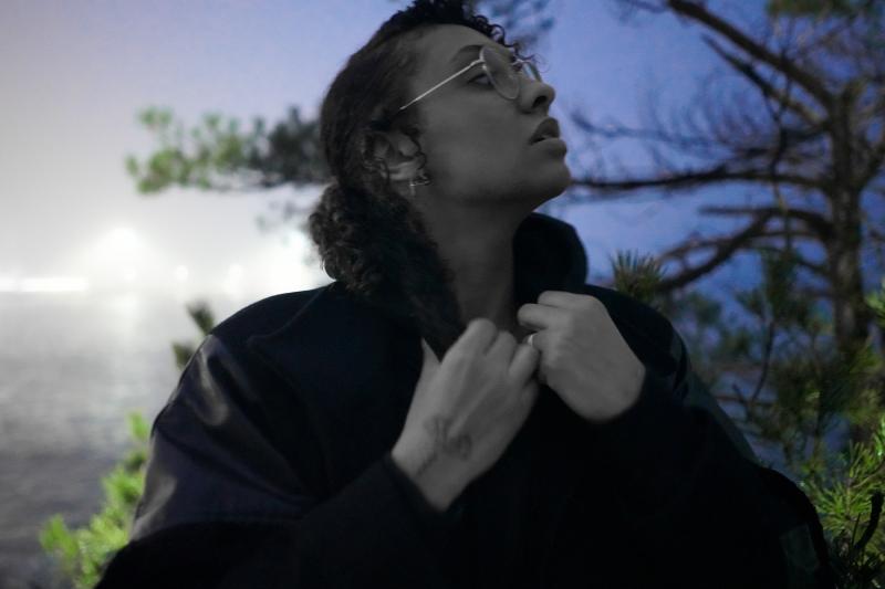 Photo of a black woman wearing glasses holding the neck of her coat, with trees and water in the background.