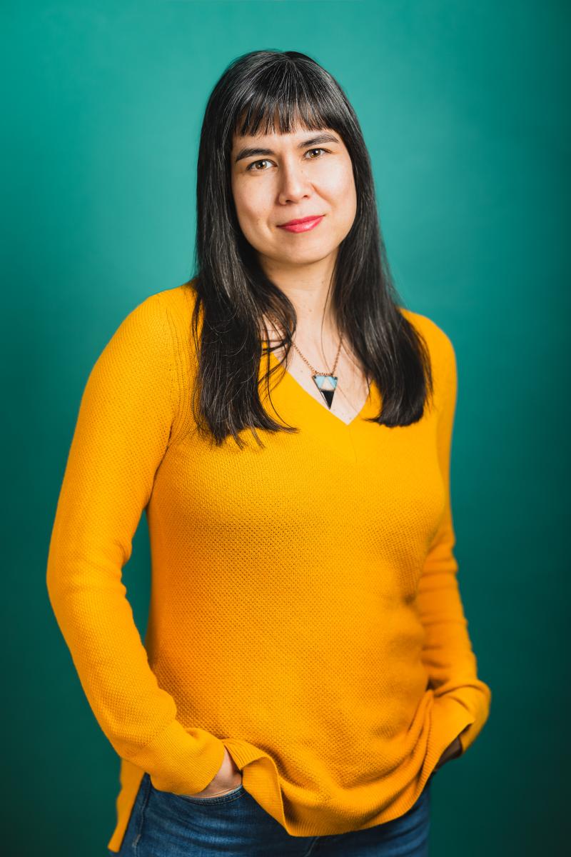 A mixed-heritage Chinese woman in her 40s with long hair, stands against a teal coloured wall in a bright yellow sweater. 