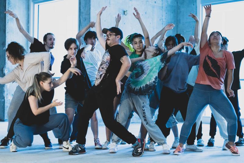 A group of people dance with their arms raised in front of a concrete wall with windows.