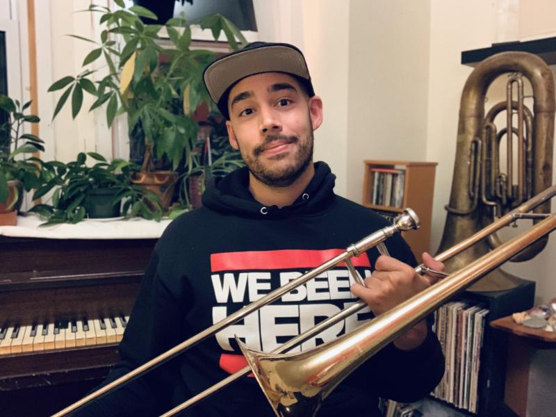 A man in a hoodie and baseball cap holds a trombone.