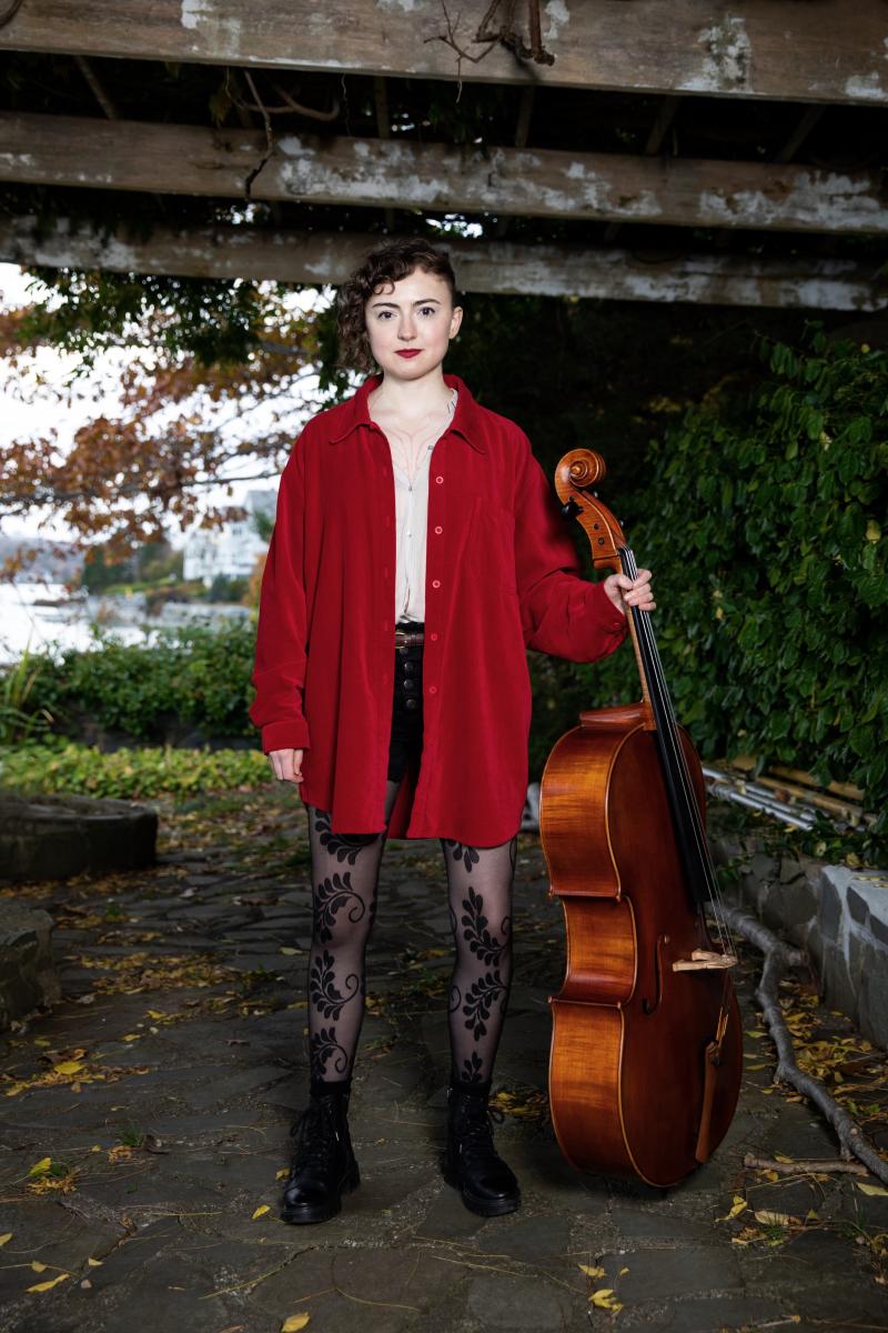A light skinned woman in a red coat stands holding a cello next to her.