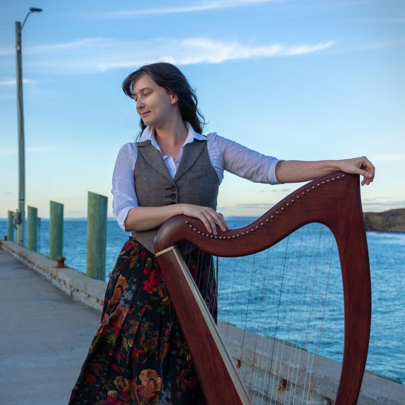 A woman in a skirt and vest stands on a pier resting her hands on a harp.