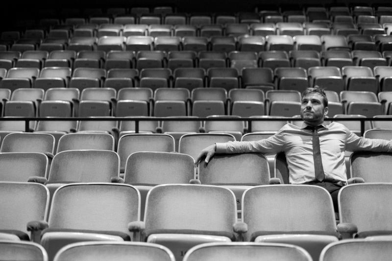 A black and white image of a man in a shirt and tie sitting in an empty theatre.