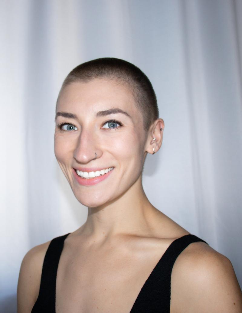 A white woman with buzzed hair smiles widely.