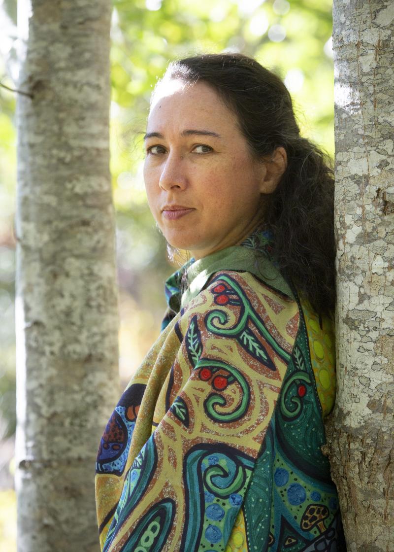 A woman in a patterned shirt stands between two trees.