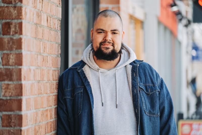 A man in a jean jacket and hoodie smiles next to a brick wall.