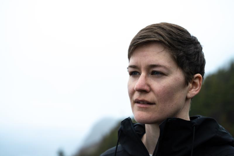 A woman stands in front of a steep forested coastline