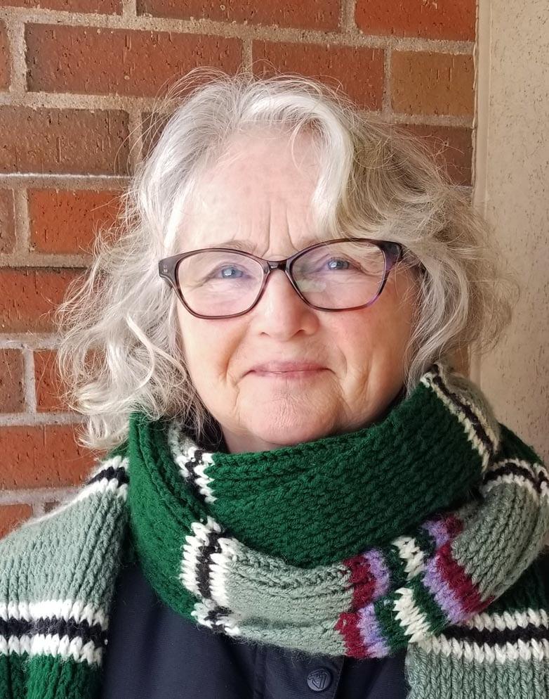 A white-haired woman in glasses and a striped scarf smiles.