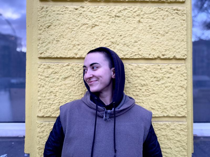 A person with short hair in a hoodie stands in front of yellow wall.