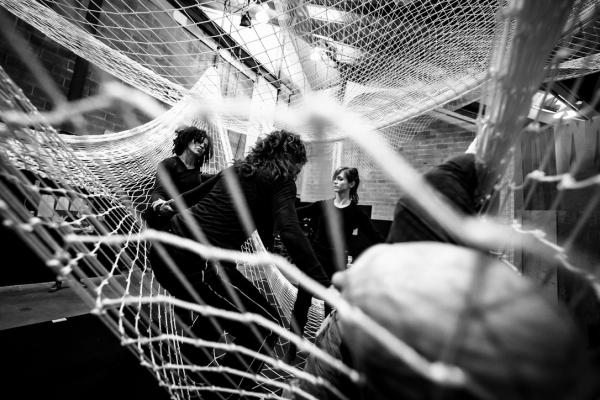 Four people sit or lay holding each others hands, within a large suspended net.