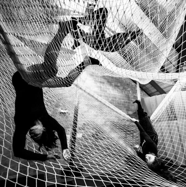Three people lie down or invert themselves within large layers of nets, suspended from the ceiling.