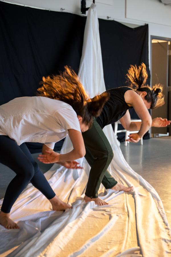 Two dancers lean forward with their arms in a cradling position, standing on a long flowing length of white fabric.