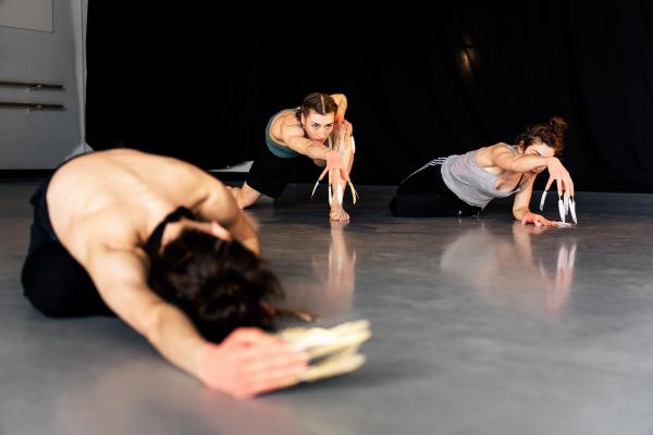 Three dancers wearing gloves with feathers on their fingers, take different positions on a white studio floor in front of a black backdrop.
