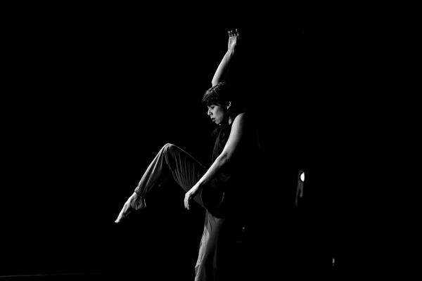 A black and white image of a dancer lifting one knee high, and one arm straight above them.