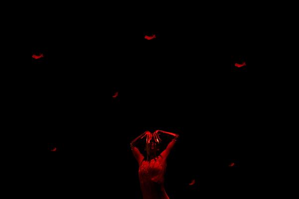 A dancer in red obscures their face behind their fingers, under a fall of red feathers.