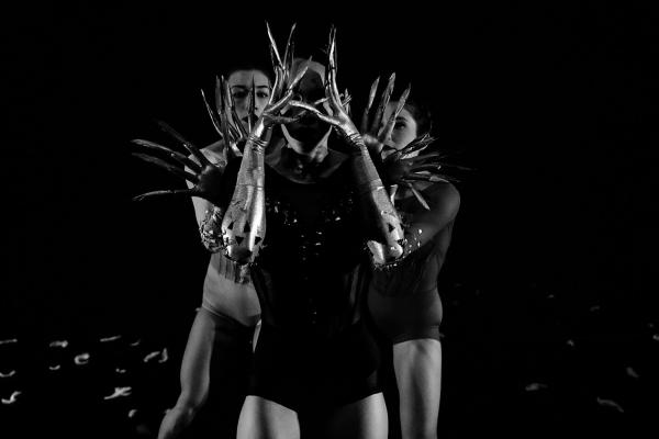 Two dancer huddle behind a third, whose face is framed and obscured by their hands, gloved in gold, with fingertips extended by feathers.