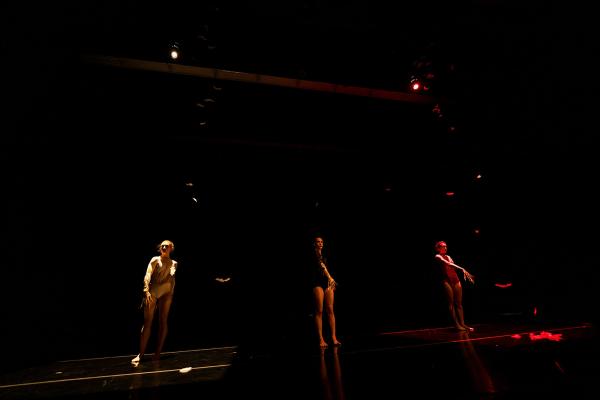 Three dancers stand in a dark space in their own spotlights.