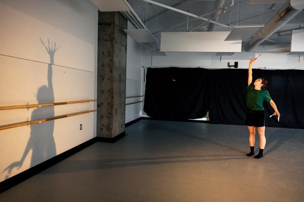 A dancer reaches one arm high, casting a large shadow on the studio wall.