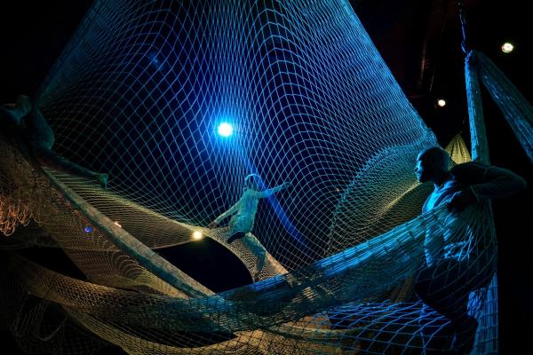 Three people spread out standing or sitting in laryers of nets suspended from the ceiling under blue lights.