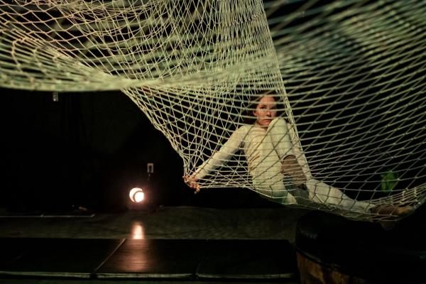 A woman in white sits with one leg bent up in an large white net, suspended over the ground.
