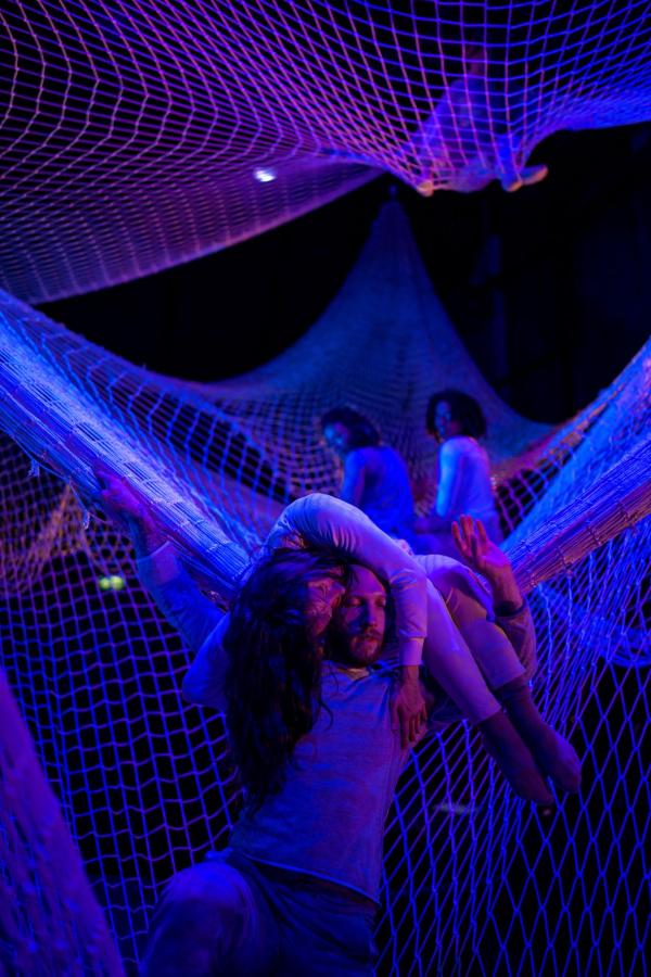 A man stands at the edge of a large suspended net, holding a woman curled up around his shoulders. Two people high up in the net look down at them, under blue and purple lights.
