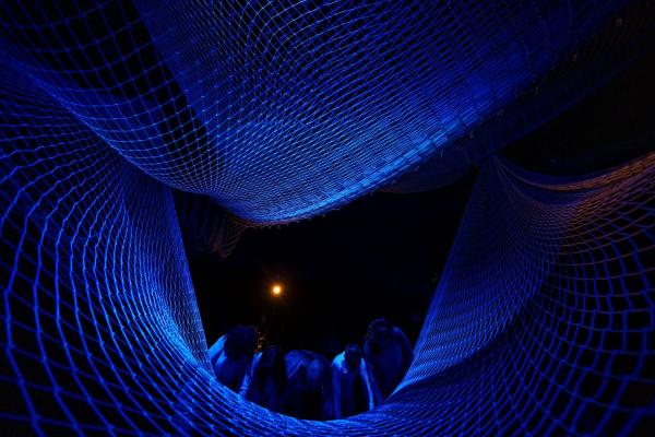 Five people push down on the edge of a suspended net, creating a curved tunnel with the layer of netting above them, all lit in shades of blue.