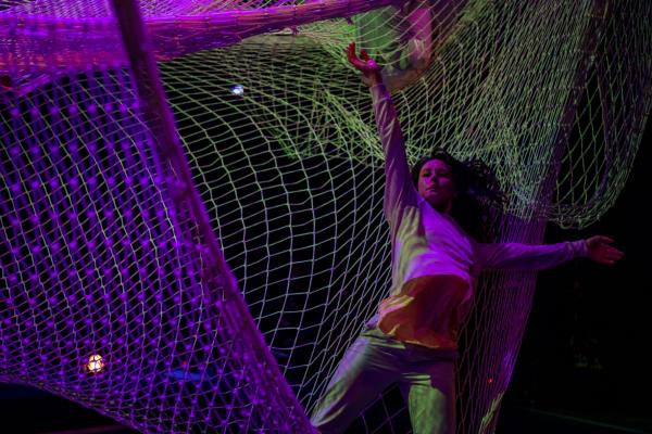 A woman stands in the edge of a web of suspended nets, one arm opening side, the other reaching high.