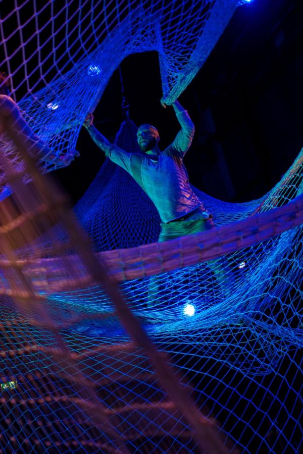 A man stands in a net suspended from the ceiling, reaching to hold the edge of another layer of netting above him, lit in shades of blue and pink.