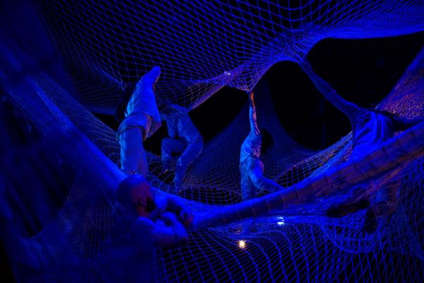 Four people stand in a large net suspended from the ceiling, reaching up to grasp another layer of netting above them. A man stands at the lower edge, looking up at the scene under blue lights.