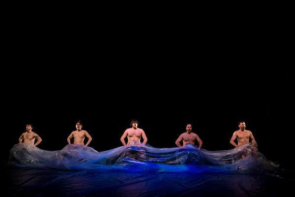 Five shirtless men sit in a line with a large plastic sheet draped over their laps.