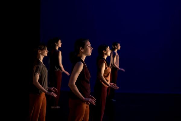Five women are spread out in a dark space. Facing the side, they step forwards with their hands raised tentatively at hip level.