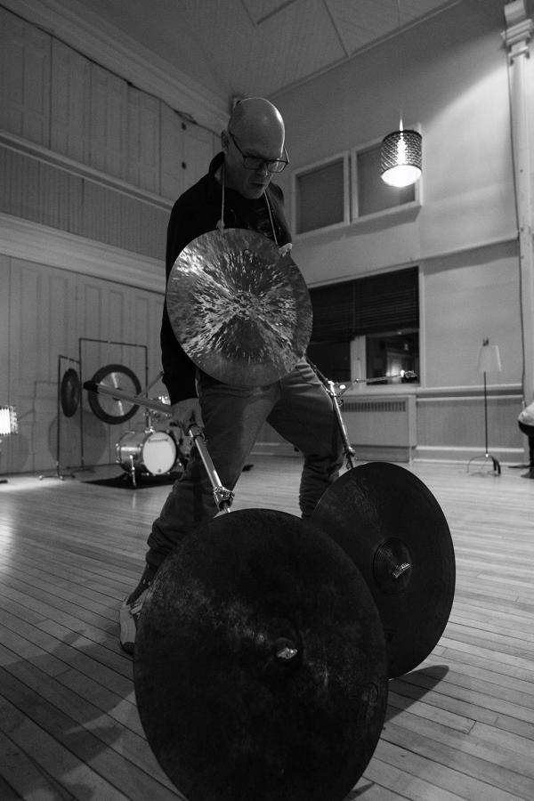 A man stands wearing a gong around his neck, balancing two cymbals on the floor on their side edge.