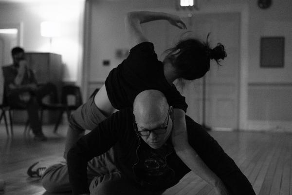 A man lies down holding his upper body off the floor and supporting a woman stretched out over his back.
