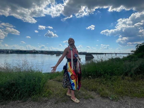 A woman in a colorful regalia stands in front of a body of water under a clouded blue sky