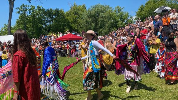 Many women in colourful regalia make their way onto a wide grassy clearing surrounded by spectators