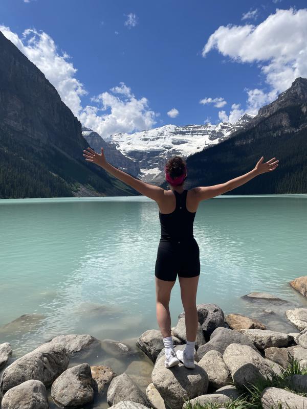 A person stands facing away with arms raised, in front of a blue lake and mountains