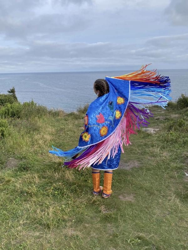 A woman stands on grass next to the ocean, swinging a colourful fringed shawl.
