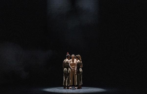 Three dancers stand in a close circle under a spotlight on a dark stage.