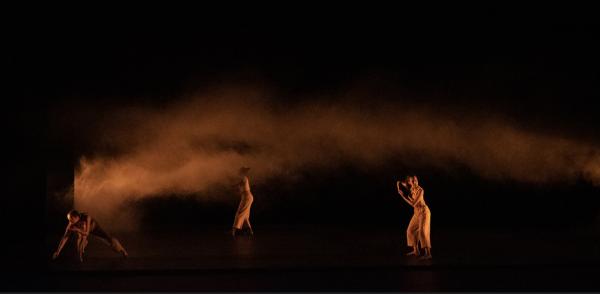 Three dancers spread out in a black space filled with smoke glowing with orange light