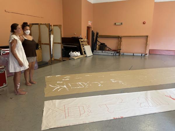 Two women stand next to long sheets of paper laid on the floor, covered in symbols.