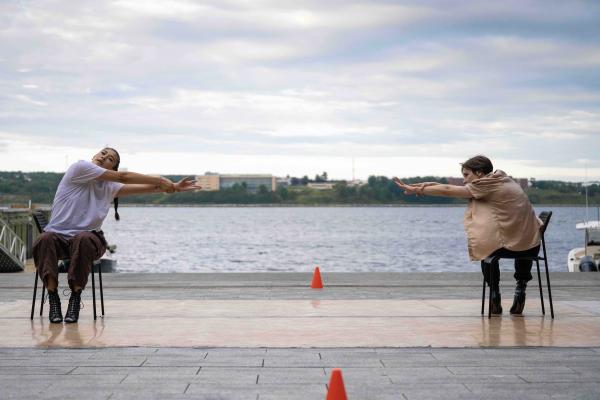 Two people dancing on chairs reach towards each other in front of a harbour