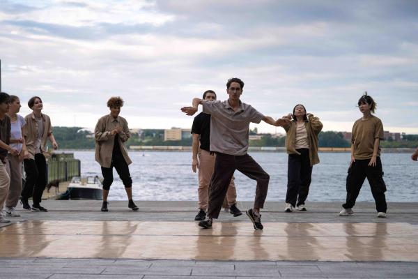 A person dances solo surrounded by a group, in front of a harbour