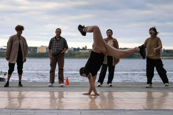 A person breakdances in front of a harbour while others look on.
